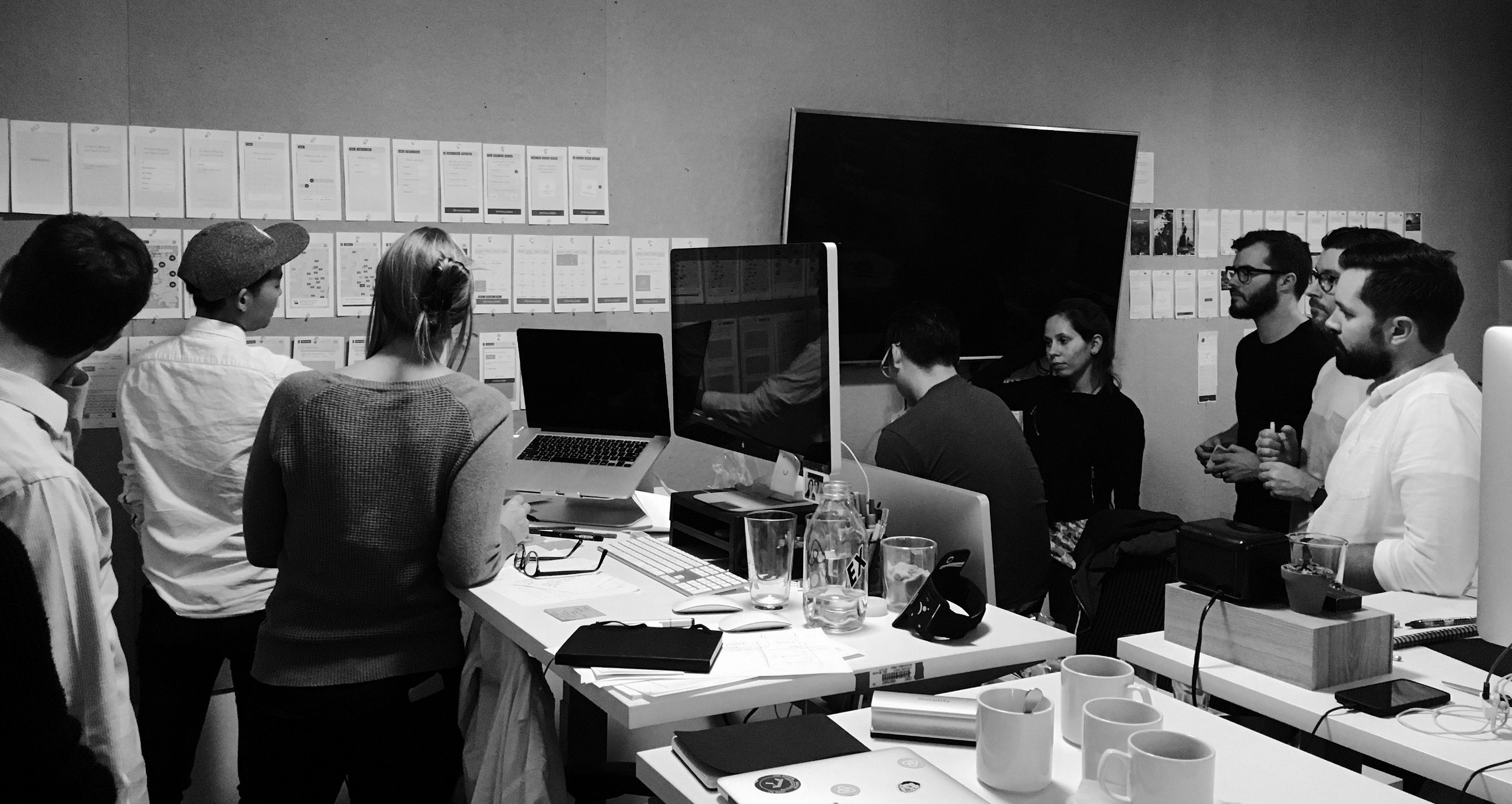 Black and white image. Katie is presenting to a group of people. Katie is standing in front of wireframes pinned up on a board.